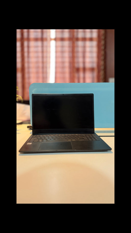 A black laptop with an open screen and keyboard, placed on a light-colored desk in front of a blue divider, with a blurred indoor background.