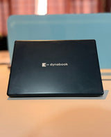 A black Dynabook laptop is shown closed, resting on a light-colored surface with a blurred blue chair and warm-toned curtains in the background.