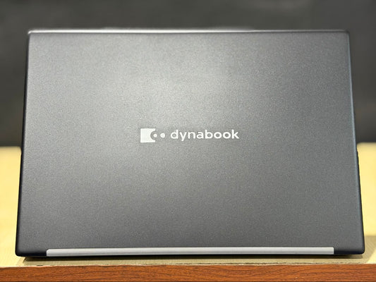 A closed Dynabook laptop shown from the back, featuring a dark gray textured lid with the white Dynabook logo in the center, resting on a wooden surface with a dark background.