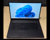 A black laptop with the screen turned on, displaying the Windows 11 desktop, placed on a light wood desk against a dark background.