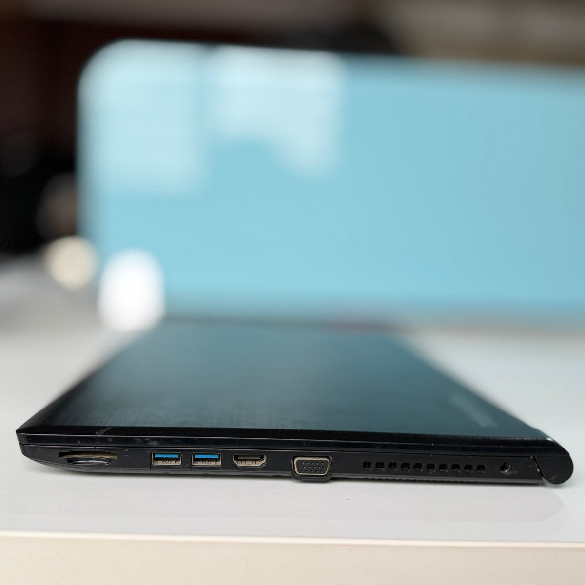 Close-up side view of a black laptop showing its ports, including USB-A, USB-C, HDMI, and VGA, resting on a white surface with a blurred light blue background.