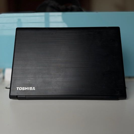 A closed black Toshiba laptop with a brushed metal finish, resting on a light gray surface with a light blue background.