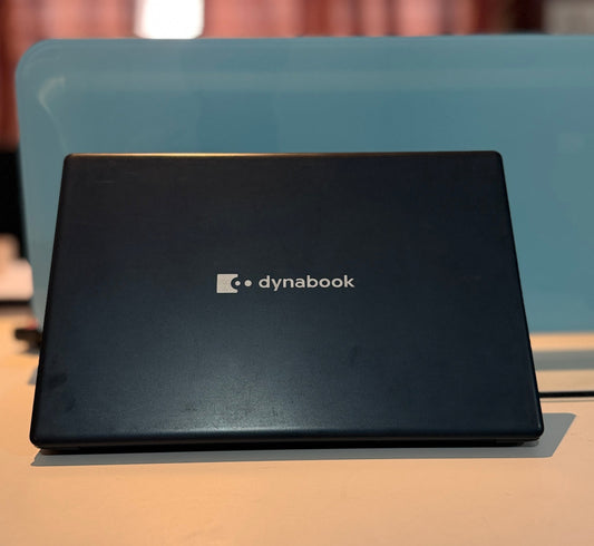 A closed black Dynabook laptop sits on a light-colored surface in front of a blue chair, with a softly blurred indoor background.
