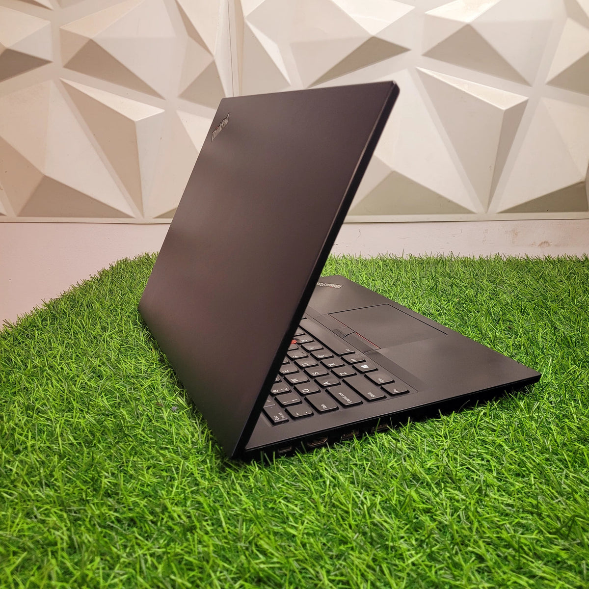 A black Lenovo ThinkPad E490 laptop placed on artificial grass against a geometric patterned backdrop.