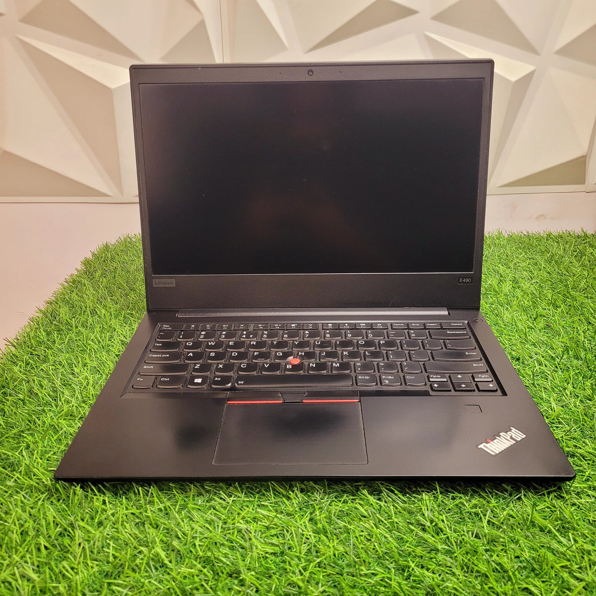 A black Lenovo ThinkPad E490 laptop with a 14-inch display, keyboard visible, placed on a grass-like surface.