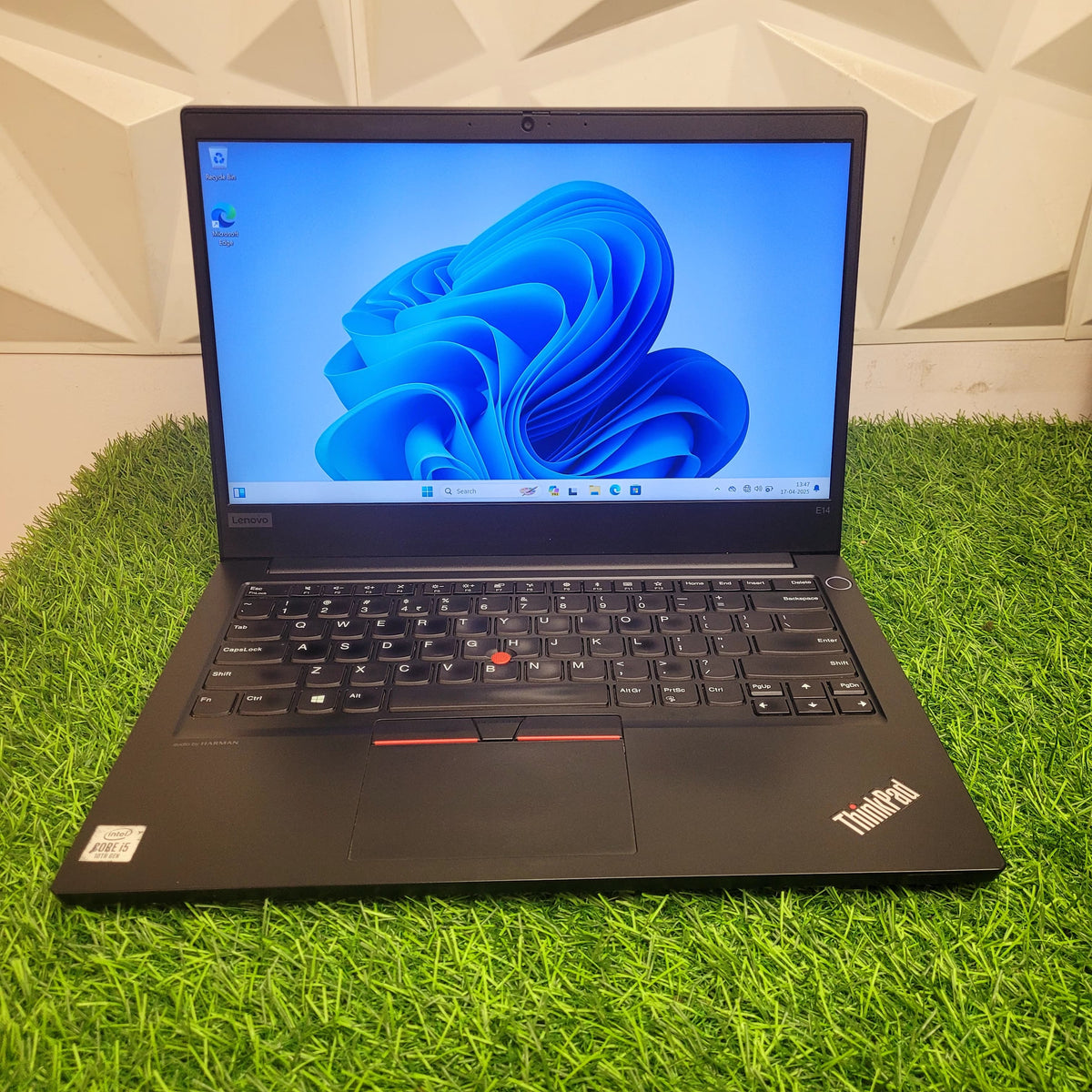A Lenovo ThinkPad E14 laptop with a black color scheme, featuring a Full HD display, visible keyboard, and trackpad on a green background.