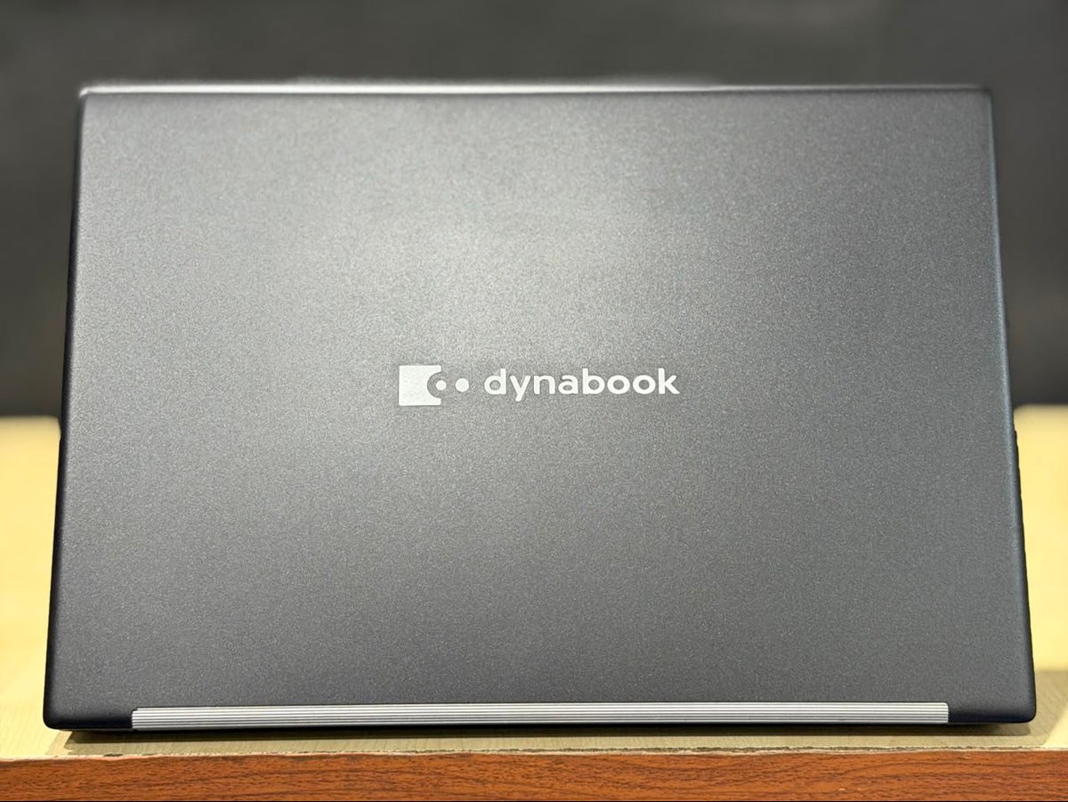 A closed Dynabook laptop shown from the back, featuring a dark gray textured lid with the white Dynabook logo in the center, resting on a wooden surface with a dark background.