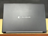 A closed Dynabook laptop shown from the back, featuring a dark gray textured lid with the white Dynabook logo in the center, resting on a wooden surface with a dark background.