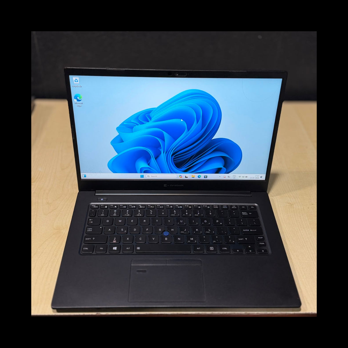 A black laptop with the screen turned on, displaying the Windows 11 desktop, placed on a light wood desk against a dark background.