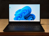 A black Microsoft Surface laptop is open and placed on a wooden table, displaying the blue Windows 11 desktop wallpaper. The background is a dark, textured wall.