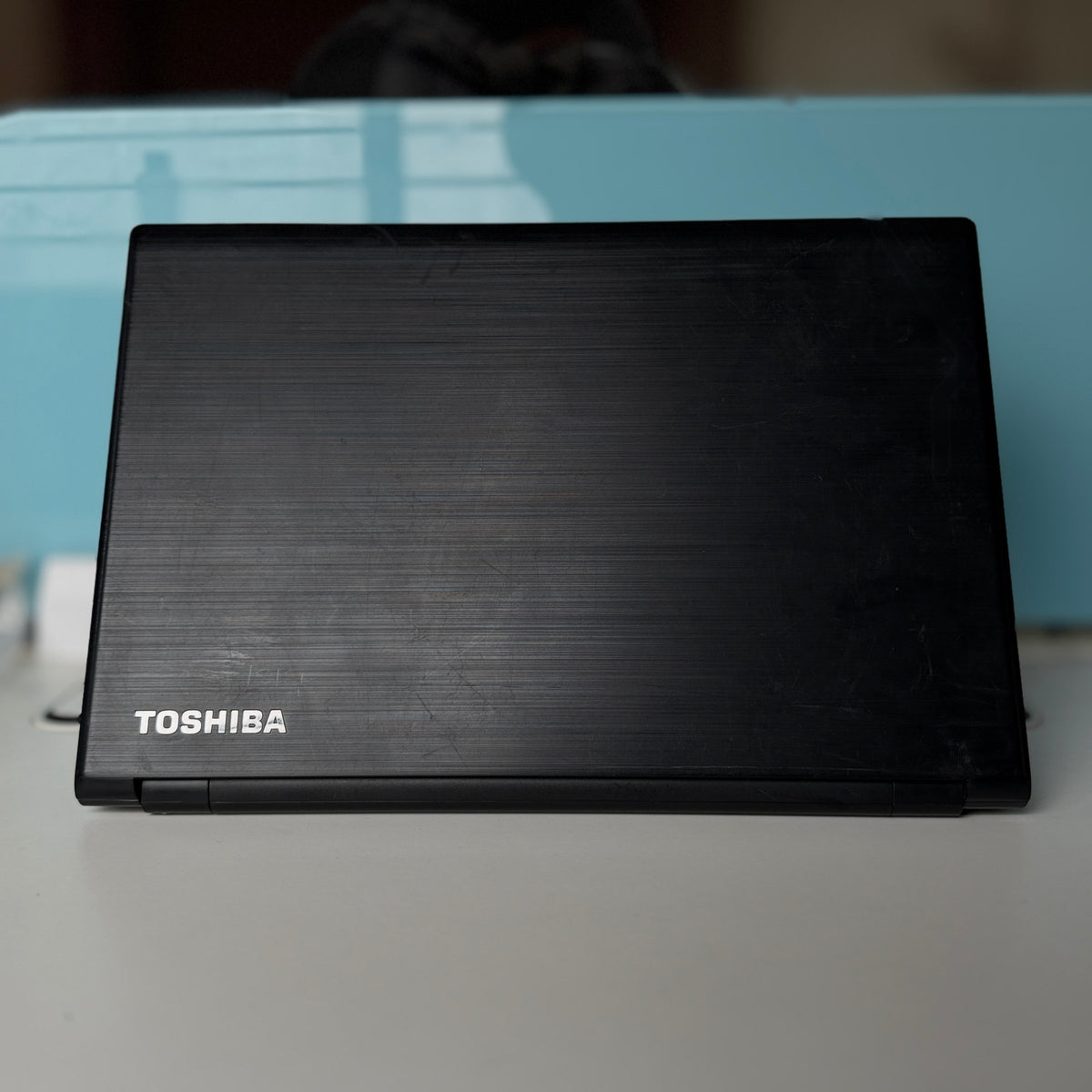 A closed black Toshiba laptop with a brushed metal finish, resting on a light gray surface with a light blue background.