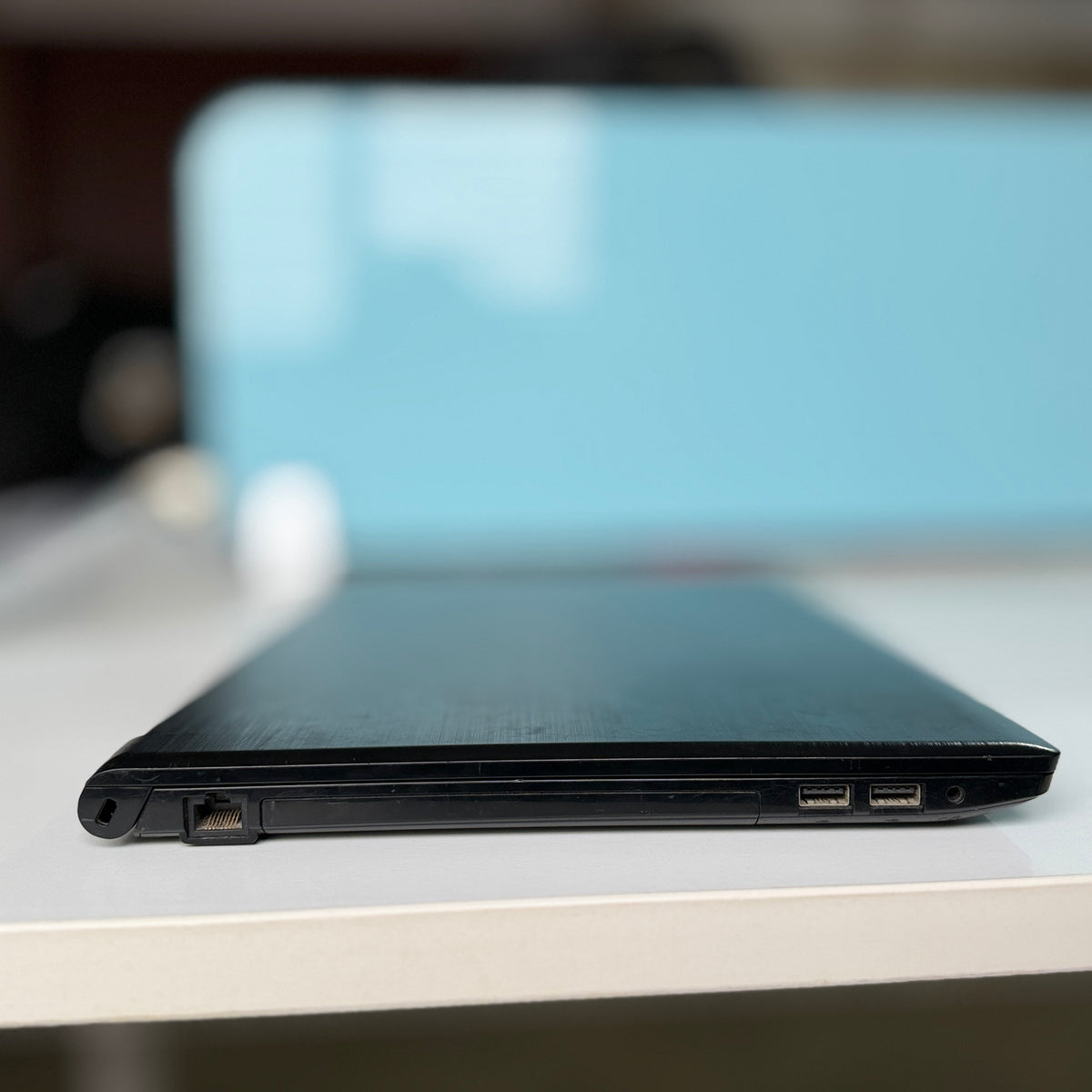 A close-up side view of a black laptop resting on a white surface, showing the thin edge of the lid and ports on the side, with a blurred light blue background.