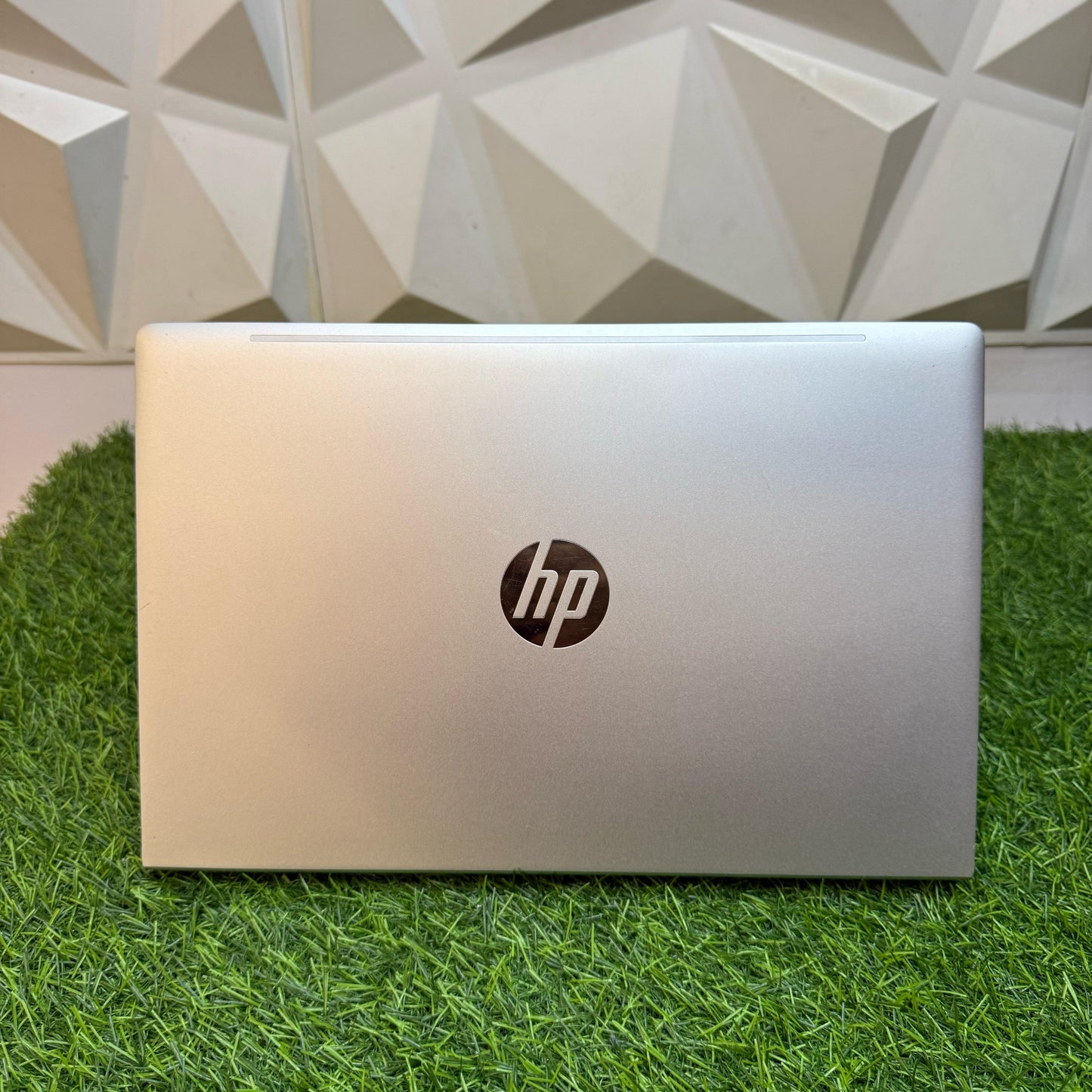 Closed silver HP ProBook 440 G8 laptop on a grass-like surface with a geometric patterned wall in the background.
